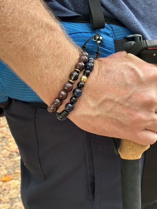 Person hiking with two beaded bracelets—dark brown and black—gripping a cork-handled pole. Earth &amp; Shadow style: grounded, resilient, and connected to nature.